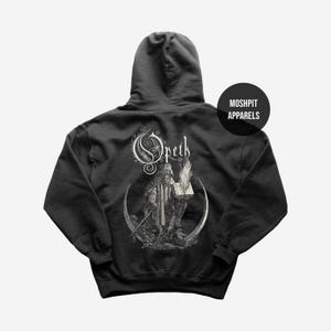 May include: Black hooded sweatshirt with a white graphic of a robed figure holding a book and a flame, and the band name "Opeth" above. The back of the sweatshirt also has the text "Moshpit Apparels" in a black circle.