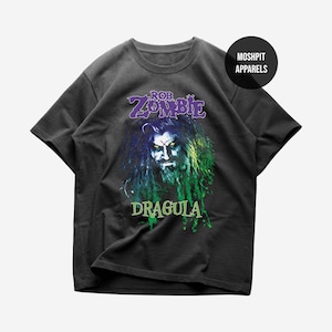 May include: Dark gray t-shirt featuring a graphic of Rob Zombie with the word "DRAGULA" in green. The text "ROB ZOMBIE" is in purple above the image. The shirt is from Moshpit Apparels.