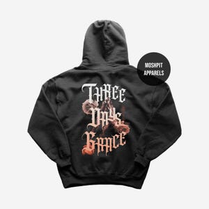 May include: Black hooded sweatshirt with the text "THREE DAYS GRACE" in a gothic font, with a floral design. The sweatshirt has a drawstring hood and a kangaroo pocket. The brand name "MOSHPIT APPARELS" is printed on a black circle.