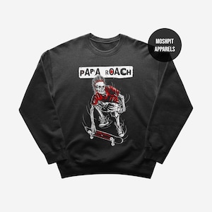 Puede incluir: Sudadera gris oscuro con un gráfico de un esqueleto patinando, con el texto "PAPA ROACH" en un rectángulo blanco. El esqueleto lleva una camisa roja. La sudadera tiene cuello redondo.