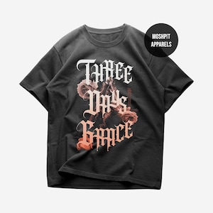 May include: Dark gray t-shirt featuring the text "Three Days Grace" in a gothic font, accompanied by a floral design with roses in shades of pink and orange. The shirt has a crew neck and short sleeves. The brand name "Moshpit Apparels" is in a black circle.