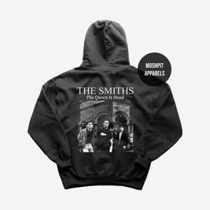 Op de afbeelding: Zwarte hoodie met een zwart-wit afbeelding van The Smiths en de tekst "THE SMITHS The Queen Is Dead." De hoodie heeft ook de tekst "MOSHPIT APPARELS" in een zwarte cirkel.