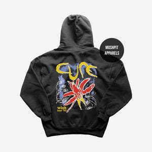 Op de afbeelding: Zwarte hoodie met een grafisch ontwerp op de rug. Het ontwerp toont het woord "Cure" in geel, een rood en gele ster en de tekst "wish tour '92". Een zwarte cirkel met de tekst "Moshpit Apparels" staat rechtsboven.