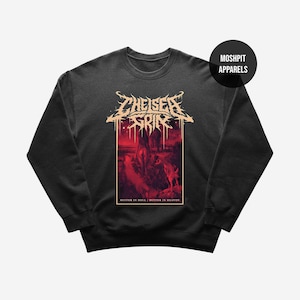 Puede incluir: Sudadera gris oscuro con un gráfico rojo y negro. El gráfico presenta el texto "CHEISER GRIN" en una fuente estilizada, una escena de paisaje y el texto "SUFFER IN HELL, SUFFER IN HEAVEN". La marca "MOSHPIT APPARELS" también es visible.