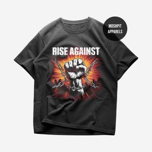 May include: Black t-shirt featuring a graphic of a clenched fist breaking chains, with the words "RISE AGAINST" in white block letters. The design includes a red and orange burst background and a cityscape silhouette.