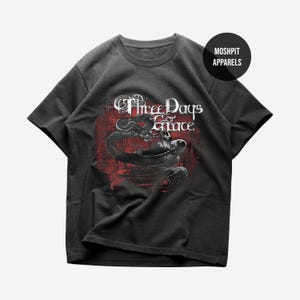 May include: Dark gray t-shirt featuring the band name "Three Days Grace" in white script above a black and white illustration of a snake on a red background. The shirt has a crew neck and short sleeves.