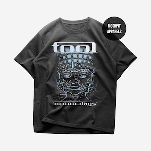 May include: Dark gray t-shirt featuring the band name "TOOL" in white block letters above a detailed illustration of a face with multiple layers and the text "10,000 days" below. The shirt has short sleeves and a crew neck.