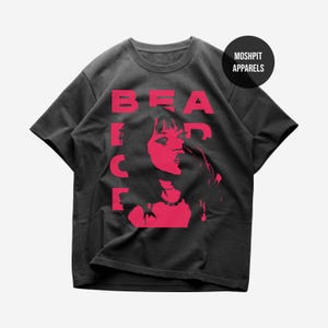 May include: Dark grey t-shirt with a red graphic. The graphic features the word "BEACH" vertically stacked with a red silhouette of a person. The brand name "MOSHPIT APPARELS" is printed on a black circle.