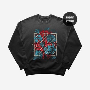 Op de afbeelding: Donkergrijze sweatshirt met een bloemenontwerp in een witte rechthoekige lijst. Het ontwerp bevat rode en blauwe bloemen. De woorden "I Prevail" en "Post Traumatic" staan boven en onder het bloemenontwerp gedrukt. Een zwarte cirkel met "Moshpit Apparels" staat rechtsboven.