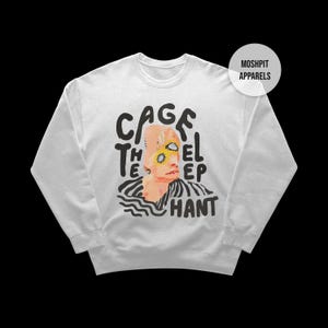 Può includere: Felpa bianca con un design grafico. Il design presenta il testo "CAGE THE EL EP HANT" in nero e un'illustrazione stilizzata di una persona con accenti gialli. Un logo circolare con "MOSHPIT APPARELS" si trova nell'angolo in alto a destra.