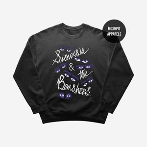 Pode incluir: Camisola preta com gola redonda com o texto branco "Siouxsie & the Banshees" e gráficos de olhos roxos. A camisola tem gola redonda e mangas compridas. Um círculo preto com o texto branco "Moshpit Apparels" está no canto superior direito.