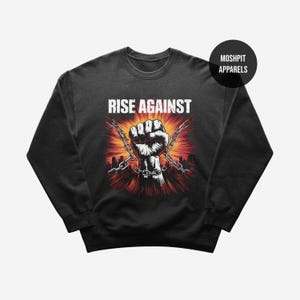 May include: Black sweatshirt with a graphic design featuring a clenched fist breaking chains, with the words "RISE AGAINST" in white block letters. The design includes a cityscape silhouette and a burst of red and orange light.