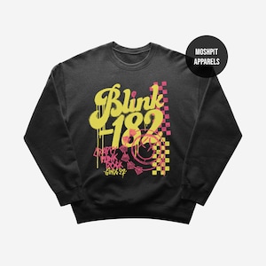 May include: Black crewneck sweatshirt with a yellow and pink graphic design. The design features the text "Blink 182" in a graffiti-style font, along with other text and a checkered pattern. The sweatshirt has a round neckline.