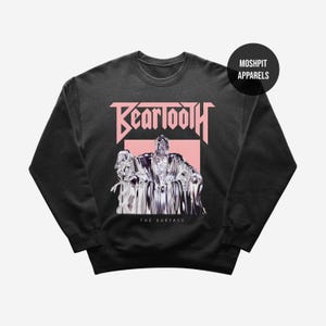 May include: Black crew neck sweatshirt with a pink Beartooth logo and a graphic of a figure. The text "THE SURFACE" is below the graphic. The sweatshirt has a round neckline and long sleeves. A black circle with "MOSHPIT APPARELS" is in the upper right.
