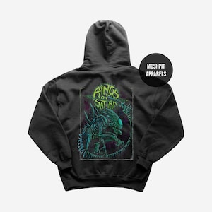 Op de afbeelding: Donkergrijze hoodie met een neon groen en blauw alien-ontwerp en de tekst "Rings of Saturn". De achterkant van de hoodie toont een gedetailleerde illustratie van een buitenaards wezen. De merknaam "Moshpit Apparels" is gedrukt op een zwarte cirkel.