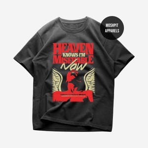 Op de afbeelding: Donkergrijs t-shirt met een rode en witte afbeelding. De afbeelding bevat de tekst "HEAVEN KNOWS I'M MISERABLE NOW" boven een rode illustratie met engelen vleugels. De tekst "Moshpit Apparels" staat in een zwarte cirkel.
