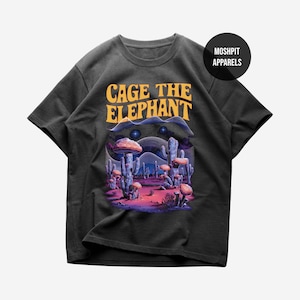 Può includere: T-shirt grigio scuro con la scritta "CAGE THE ELEPHANT" in arancione e una grafica colorata di un paesaggio desertico. La grafica include cactus, funghi e una combinazione di colori viola e rosa. La maglietta ha un girocollo e maniche corte.