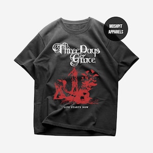 May include: Black t-shirt featuring the band name "Three Days Grace" in white script above a red graphic design. The text "LIFE STARTS NOW" is printed in red below the graphic. The shirt has a crew neck and short sleeves.