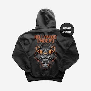 May include: A black hooded sweatshirt featuring a wolf graphic with fiery orange eyes and accents. The text "HOLLYWOOD UNDEAD" is printed above the wolf's head in orange. The brand name "MOSHPIT APPARELS" is printed on a black circle on the upper right.