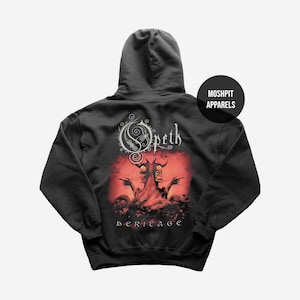 Puede incluir: Sudadera con capucha negra con el logotipo de la banda Opeth y la ilustración del álbum. El diseño incluye un fondo rojo con dos figuras y la palabra "heritage" debajo. La sudadera tiene una capucha con cordón y un logotipo de "Moshpit Apparels".