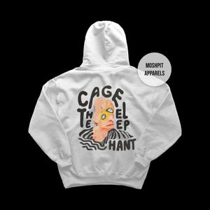 Può includere: Una felpa con cappuccio grigio chiaro con una grafica sul retro. La grafica presenta un volto stilizzato con dettagli gialli e neri e la scritta "CAGE THE ELEPHANT". La felpa ha anche la scritta "MOSHPIT APPARELS".