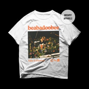 May include: White t-shirt with the orange text "beabadoobee" above a photo of a person playing a guitar amidst flowers. The words "Fake It Flowers" are below the photo, with the "Moshpit Apparels" logo in the upper right.