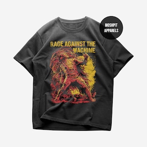 Puede incluir: Camiseta gris oscuro con un gráfico de una figura en tonos rojos y amarillos, con el texto "RAGE AGAINST THE MACHINE" en amarillo. La camiseta tiene mangas cortas y cuello redondo, con el logo "Moshpit Apparels" en la esquina superior derecha.