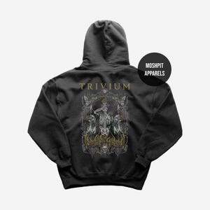 May include: Dark gray hoodie featuring a detailed graphic of skeletons, flames, and ornate framing. The word "TRIVIUM" is printed across the upper back in gold lettering. A black circle with "MOSHPIT APPARELS" is on the upper right.