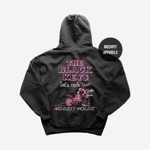 Può includere: Felpa con cappuccio nera con testo rosa "THE BLACK KEYS 'Let's rock' tour MODEST MOUSE". Il retro della felpa presenta una grafica di una band che suona strumenti. Un cerchio nero con testo bianco "MOSHPIT APPARELS" è in alto a destra.