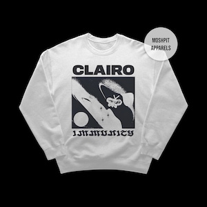 May include: White sweatshirt with the word "CLAIRO" above a black and white graphic. The graphic includes a stylized figure, a circle, and stars. The word "IMMUNITY" is printed below. A "MOSHPIT APPARELS" logo is on the upper right.