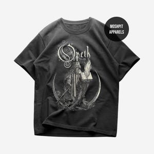 Puede incluir: Camiseta gris oscuro con una ilustración en blanco y negro de una figura vestida con una túnica que sostiene un libro y una antorcha en llamas, sobre una media luna. La palabra "Opeth" se muestra en una fuente adornada. La camiseta tiene cuello redondo y mangas cortas.