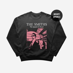 Op de afbeelding: Donkergrijze sweatshirt met een roze afbeelding van twee mannen en de tekst "THE SMITHS The Queen Is Dead". De sweatshirt heeft ook de tekst "MOSHPIT APPARELS" in een zwarte cirkel.