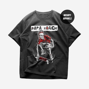 Puede incluir: Camiseta gris oscuro con un gráfico rojo y blanco de un esqueleto patinando. El esqueleto lleva una camisa roja y tiene una cresta mohawk. El texto "PAPA ROACH" está encima del gráfico en un rectángulo blanco.