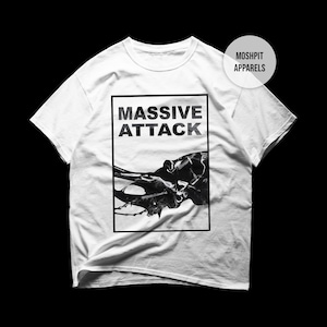 Könnte beinhalten: Weißes T-Shirt mit einem schwarz-weißen Grafikdesign. Das Design zeigt den Text "MASSIVE ATTACK" in fetter Schrift, über einem abstrakten Bild. Das T-Shirt hat einen Rundhalsausschnitt und kurze Ärmel, ein gängiger Stil für Freizeitkleidung.
