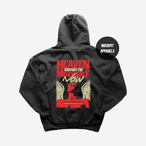 Op de afbeelding: Zwarte hoodie met de tekst "HEAVEN KNOWS I'M MISERABLE NOW" in rood en wit. Een rode afbeelding van een figuur met vleugels staat onder de tekst. De merknaam "MOSHPIT APPARELS" staat op een zwarte cirkel.