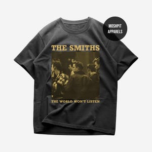 Op de afbeelding: Donkergrijs t-shirt met een sepia-kleurige afbeelding van een menigte. De tekst "THE SMITHS" staat boven de afbeelding en "THE WORLD WON'T LISTEN" eronder. Het shirt heeft een ronde hals en korte mouwen.