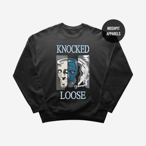 Puede incluir: Sudadera gris oscuro con el texto "KNOCKED LOOSE" en blanco, sobre un gráfico de tres caras en un panel vertical. Las caras son en tonos de blanco, azul y gris. La marca "MOSHPIT APPARELS" está en un círculo negro.