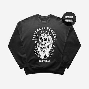 May include: Black crew neck sweatshirt with a white graphic. The graphic includes a skull with horns, hands, and the text "FALLING IN REVERSE" and "LAS VEGAS". The sweatshirt has a round neckline and long sleeves. The brand name "MOSHPIT APPARELS" is in a black circle.