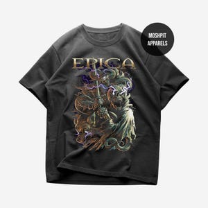 May include: Dark gray t-shirt featuring a detailed graphic design. The design includes the word "ERICA" in gold lettering above a skeletal figure holding a staff, with a wolf and lightning bolts. The shirt has a crew neck and short sleeves.