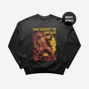 May include: Dark gray sweatshirt featuring a graphic of a figure with red and yellow accents, with the text "RAGE AGAINST THE MACHINE" in yellow. The sweatshirt has a crew neck and long sleeves. The brand name "MOSHPIT APPARELS" is in a black circle.