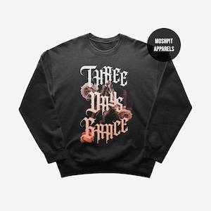 May include: Black crewneck sweatshirt with the text "Three Days Grace" in a gothic font, accompanied by floral and guitar graphics. The sweatshirt has a round neckline and long sleeves. The brand name "Moshpit Apparels" is in a black circle.