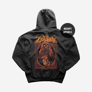 May include: Dark gray hoodie featuring a graphic of Rob Zombie with long orange hair and a skull. The design is in orange and red tones, with the text "ROB ZOMBIE" above the image. The hoodie has a hood and the text "MOSHPIT APPARELS" on the upper right.