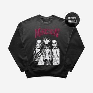 May include: Black crewneck sweatshirt featuring a black and white image of the band Måneskin. The band's name is printed in pink above the image. The sweatshirt has a round neckline and long sleeves. The brand name "Moshpit Apparels" is printed on a black circle.
