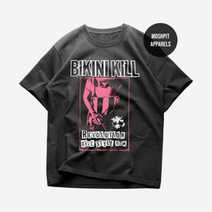 Może przedstawiać: Ciemnoszara koszulka z różową grafiką. Tekst "BIKINI KILL" znajduje się nad ilustracją, a pod nim "REVOLUTION girl STYLE now". Okrągłe logo z napisem "MOSHPIT APPARELS" znajduje się w prawym górnym rogu.
