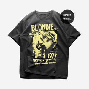 Könnte beinhalten: Dunkelgraues T-Shirt mit gelber Grafik von Blondie, dem Bandnamen und dem Jahr "1977". Das Shirt enthält auch den Text "in Los Angeles" und "Direct from New York City". Ein rundes Logo mit dem Text "Moshpit Apparels" befindet sich oben rechts.