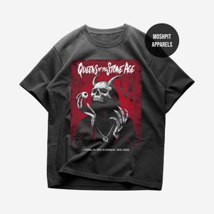 Puede incluir: Camiseta gris oscuro con un gráfico de una calavera con cuernos sosteniendo un globo ocular, sobre un fondo rojo. El texto "QUEENS OF THE STONE AGE" está encima del gráfico. La camiseta tiene mangas cortas y cuello redondo.