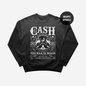 May include: Black crewneck sweatshirt with a white graphic featuring the name "CASH" and the text "THE MAN IN BLACK". The design includes guitars, stars, and the words "ORIGINAL COUNTRY ROCK". The brand name "MOSHPIT APPARELS" is also visible.