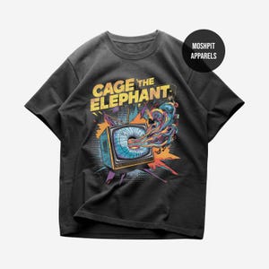 Può includere: T-shirt grigio scuro con una grafica colorata di una televisione retrò e le parole "CAGE THE ELEPHANT". Lo schermo della televisione ha un design a spirale con colori vivaci. La maglietta ha un girocollo e maniche corte. Il marchio "MOSHPIT APPARELS" è in un cerchio nero.