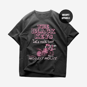 Può includere: T-shirt grigio scuro con la scritta rosa "THE BLACK KEYS 'Let's rock' tour" e un'illustrazione rosa di una band. Il testo "MODEST MOUSE" è sotto. Un cerchio nero con "MOSHPIT APPARELS" è in alto a destra.