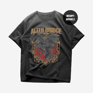 May include: Dark gray t-shirt featuring the band name "ALTER BRIDGE" in gold lettering above a detailed graphic. The graphic includes a black bird, red roses, and a snake within a decorative frame. The shirt also has the text "MOSHPIT APPARELS".